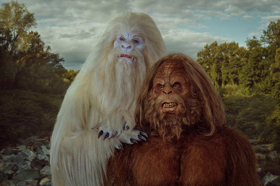 Two large, hairy creatures, one white and one brown, stand together in a wooded area with a stream, creating a mythical scene.